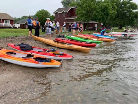 Colorful kayaks lined on a lakeshore as people in life jackets gather by cabins under cloudy skies.