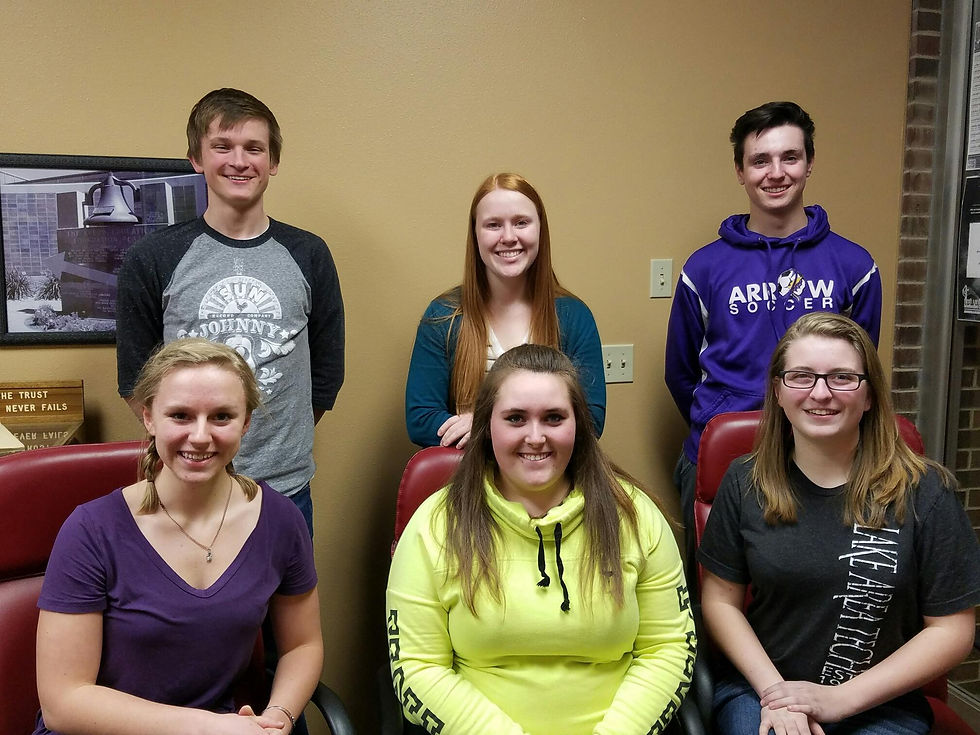 Six smiling young adults pose indoors against a tan wall; one wears a neon yellow hoodie and another a purple soccer sweatshirt.