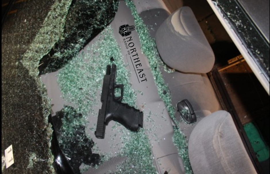 Shattered car windshield with a handgun on the front seats and Northeast Radio logo visible, eerie nighttime scene