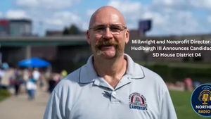 Man in glasses and gray shirt with union logo smiles outdoors. Text announces candidacy for SD House District 04. Blurred crowd behind.