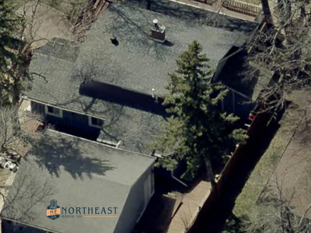 Aerial view of a house with gray roofs, tall evergreen trees, and a wooden walkway in a wooded yard; NORTHEAST RADIO SD watermark.