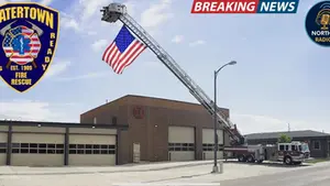 Fire truck with a long ladder displaying an American flag outside a brick fire station on a sunny day. Watertown Fire Rescue logo visible.