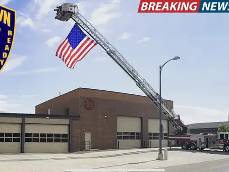 Fire truck with extended ladder displaying a US flag outside a brick fire station. Watertown Fire Rescue emblem and "Breaking News" banner visible.