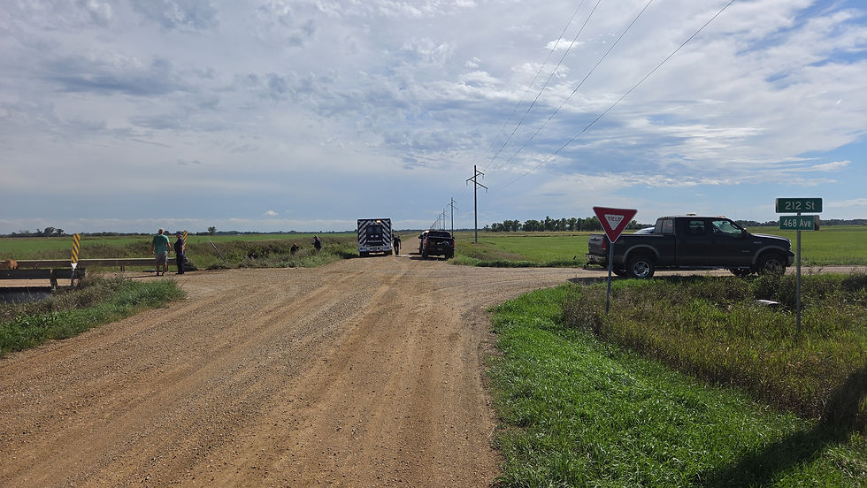 Rural dirt-road intersection with an ambulance, pickup trucks, and people near a Yield sign and 212 St/468 Ave signs under cloudy skies.