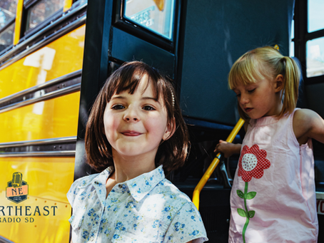 Smiling girl in floral shirt stands by yellow school bus; blonde girl in pink flower dress steps down inside. Northeast Radio SD logo.