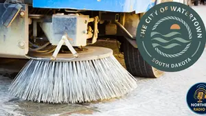 Street sweeper brush on wet pavement, blue vehicle visible. Logos: "The City of Watertown South Dakota" and "Northeast Radio SD".