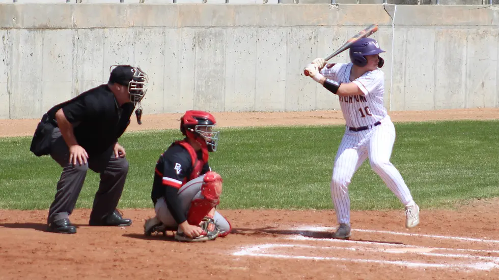 Baseball game with a batter in a white uniform, a catcher in red gear, and an umpire in black on a sunny field. Bat held ready.