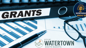 Document binder labeled "GRANTS," with charts, glasses, and a pen. Logos of Northeast Radio SD and City of Watertown, South Dakota.