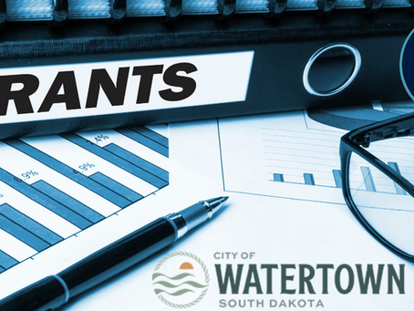 Document binder labeled "GRANTS," with charts, glasses, and a pen. Logos of Northeast Radio SD and City of Watertown, South Dakota.