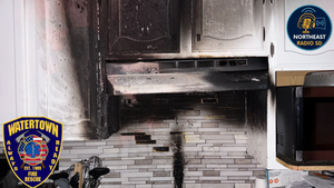 A burned kitchen cabinet and stovetop show signs of fire damage. Watertown Fire Rescue badge is visible. The mood is one of aftermath.