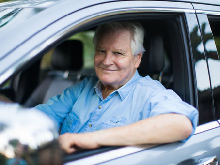 Smiling older man in a light blue shirt sits in the driver’s seat of a silver car, looking out the open window.