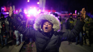 Person in fur-trimmed hooded jacket, arms raised, shouting at night. Police lights in background, with a crowd and tense atmosphere.