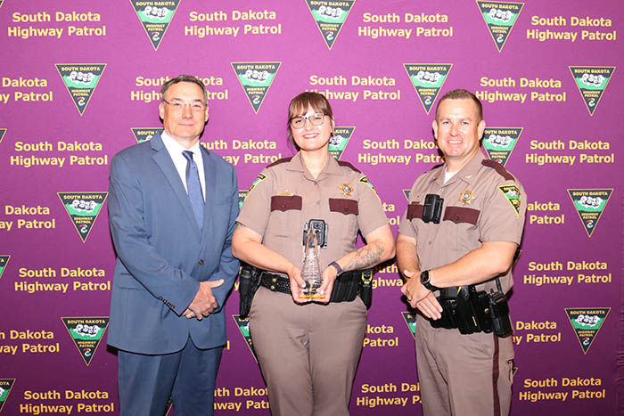 Three South Dakota Highway Patrol members pose with a clear trophy before a purple backdrop repeating South Dakota Highway Patrol.