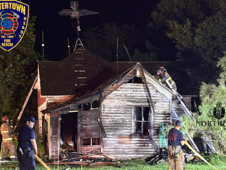 Firefighters work around a badly charred house at night, with ladders and hoses; Watertown Fire Rescue logo visible.