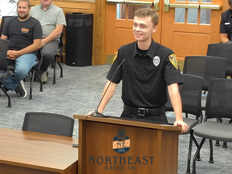 Young man in black uniform speaks at a podium labeled NORTHEAST RADIO SD in a meeting room; seated attendees watch.