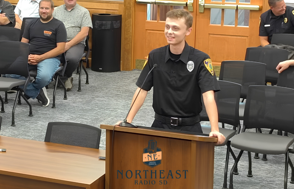 Young man in black uniform speaks at a podium labeled NORTHEAST RADIO SD in a meeting room; seated attendees watch.