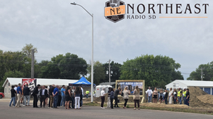 Crowd gathered at an outdoor event near a sign reading LAKE AREA LOFTS, with a Northeast Radio SD logo over a cloudy sky.