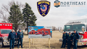 Firefighters stand beside red trucks by a Watertown Fire Rescue sign for Future Home of Station 3; Northeast Radio SD logo above.