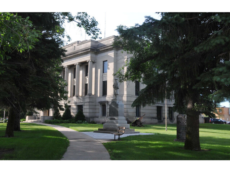 Stone courthouse with columns behind trees and lawn, a curved sidewalk and monument in front on a sunny day.