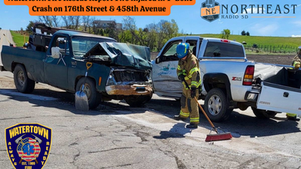 Two damaged trucks in a T-bone crash. A firefighter sweeps debris on a rural road. Text about injuries. Logos of Watertown Fire Rescue and Northeast Radio SD.