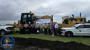Group of people, including police officers, stand in front of heavy machinery and sheriff cars on grass. Banner reads "Codington County Jail."