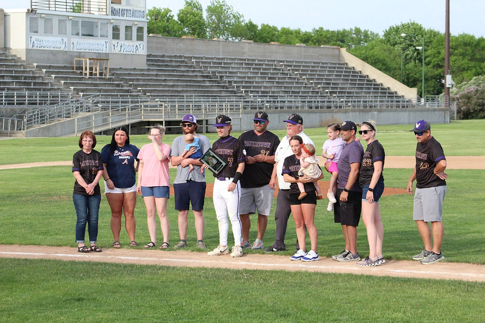 Group of Watertown baseball players and families pose on the field for a ceremony, with empty bleachers and Home of the Arrows behind.