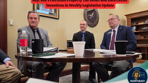 Three men in suits sit around a table with papers and cups, discussing a legislative update. A clock and framed picture are on the beige wall.