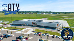 Aerial view of Watertown Regional Airport with parked cars, green fields, and clear skies. Text: "FLY ATY," "NORTHEAST RADIO SD."