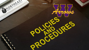 Black spiral notebook labeled POLICIES AND PROCEDURES with W Arrows logo on a desk beside a pen and Northeast Radio SD notepad.