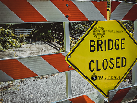 Yellow Bridge Closed sign on red-and-white barricade blocks a rural road by an overgrown bridge.