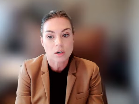 Woman in tan blazer on a video call against a blurred office background, with a small inset of another speaker in the corner.