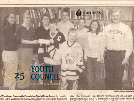 Group of youth and adults pose in a library holding a check, with Youth Council 25 Years of Youth Leadership text.
