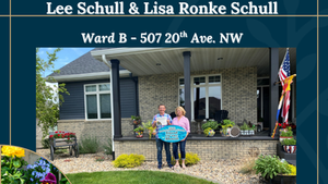 Two people hold a Yard of the Month sign on a porch of a brick house with flowers; title reads Yard of the Month
