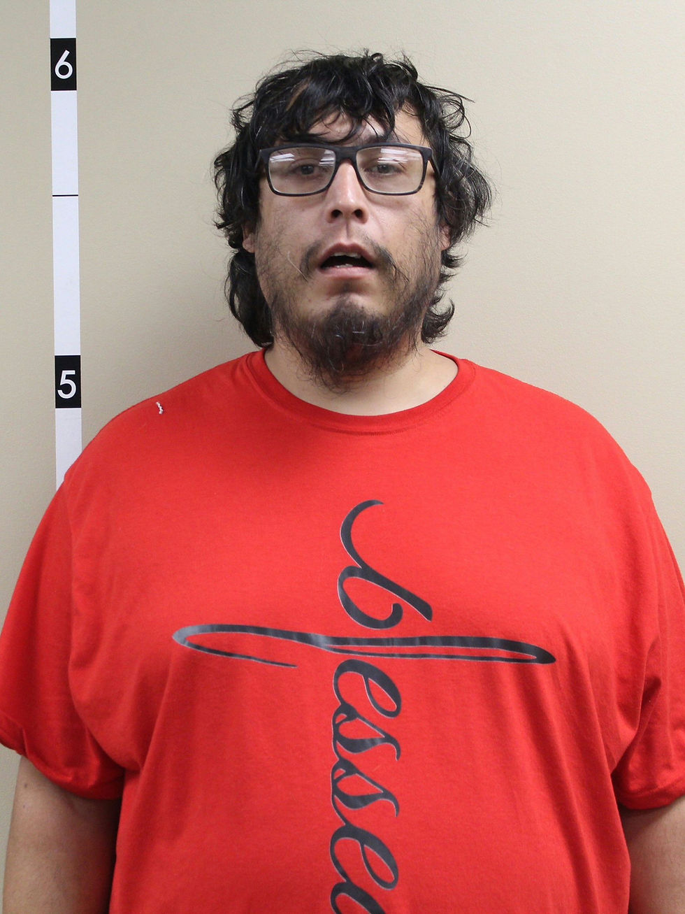 Mugshot-style photo of a man in glasses and a red Blessed T-shirt, standing against a beige wall with height markers 5 and 6.