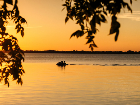 Sunset over a calm lake, silhouetted leaves framing a small boat with people gliding across golden water.