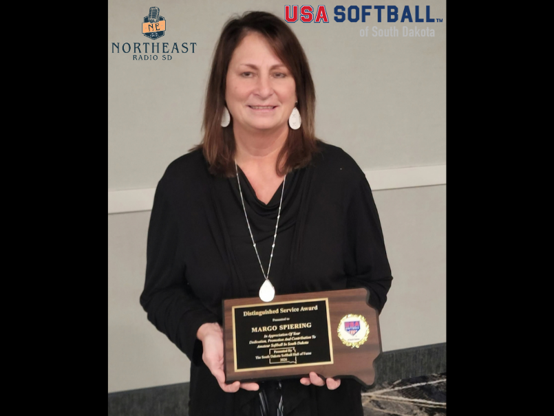Woman in black holds a Distinguished Service Award plaque, smiling before a Northeast Radio SD and USA Softball backdrop.