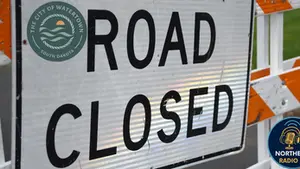 A "Road Closed" sign with orange stripes, featuring the Watertown SD and Northeast Radio SD logos, set against a blurred background.