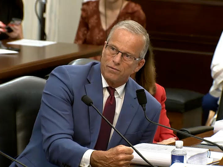 Gray-haired man in blue suit speaks at a hearing desk with microphones and papers, others seated behind him.