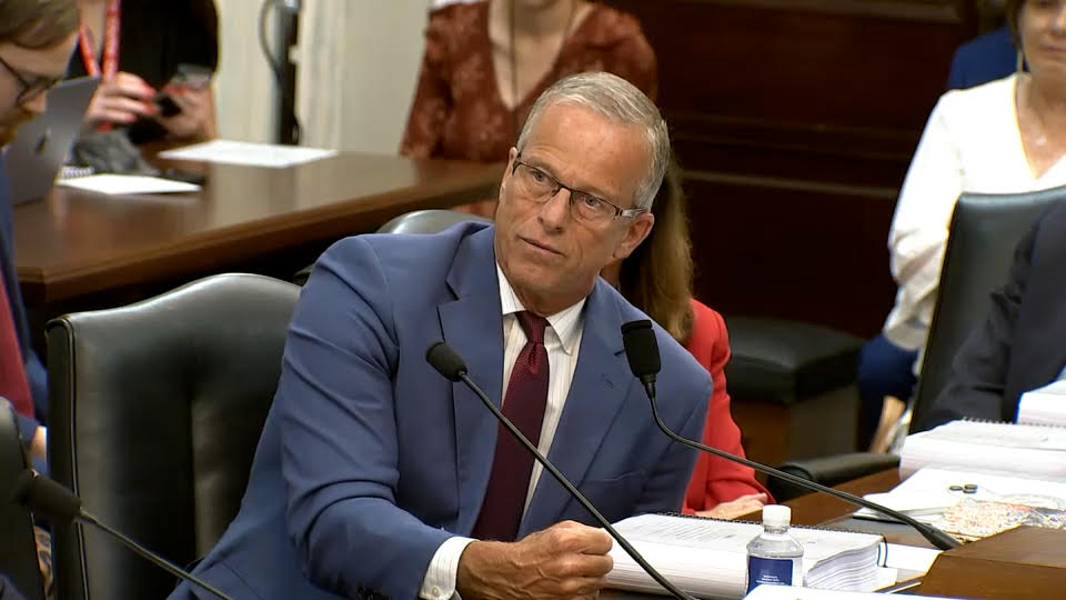 Gray-haired man in blue suit speaks at a hearing desk with microphones and papers, others seated behind him.