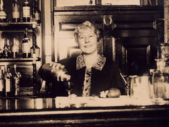 A woman stands behind a pub bar pouring a drink from a cocktail shaker