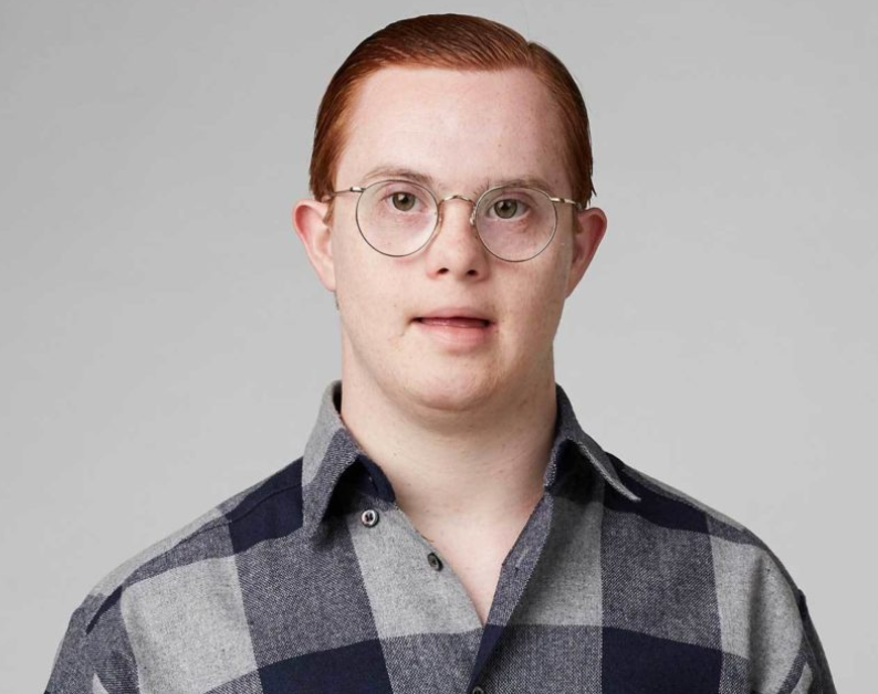 Photograph of a young ginger man with downs syndrome, wearing glasses and a grey check shirt.