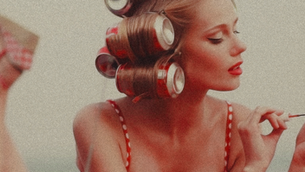 Young woman with Coke can rollers, red lipstick, and polka dot straps applies nail polish. Relaxing mood with a neutral background.