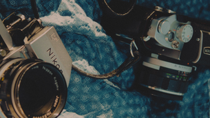Two vintage cameras, Nikon and Miranda, rest on a blue textured cloth in a moody close-up.
