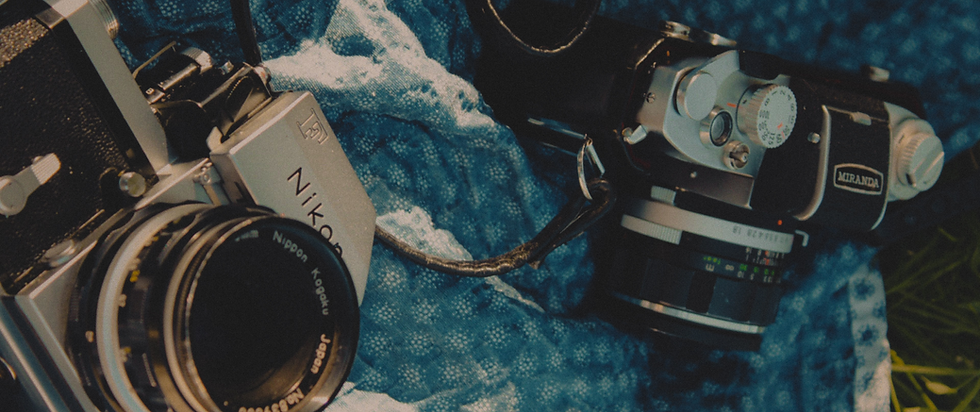 Two vintage cameras, Nikon and Miranda, rest on a blue textured cloth in a moody close-up.