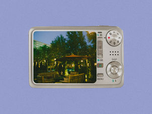 Compact camera screen shows a lit evening garden patio with trees and umbrellas; DISP and MENU buttons on the silver body.