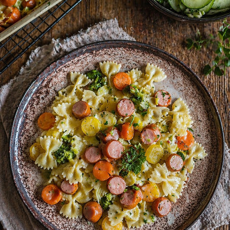 Kunterbuntes Feierabendblech mit Nudeln, Wiener Würstchen und gemischtem Gemüse in cremiger Soße, frisch aus dem Ofen und mit Kräutern garniert