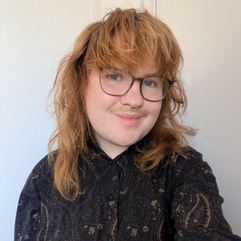 Lukas has wavy strawberry blond hair with a choppy fringe and a neat moustache.  He wears glasses, and a gold septum ring.  