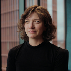 Lena has short wavy chestnut hair and wears a black jumper