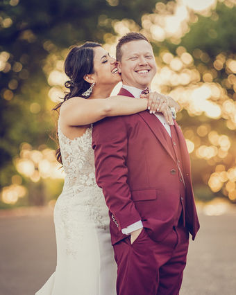 A bride hugs her groom from behind and nibbles on his ear.