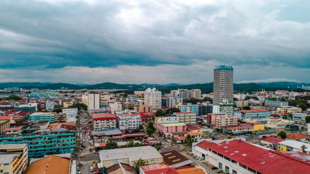 Aerial view of a colorful city skyline with a tall tower under dark, cloudy skies and distant hills.
